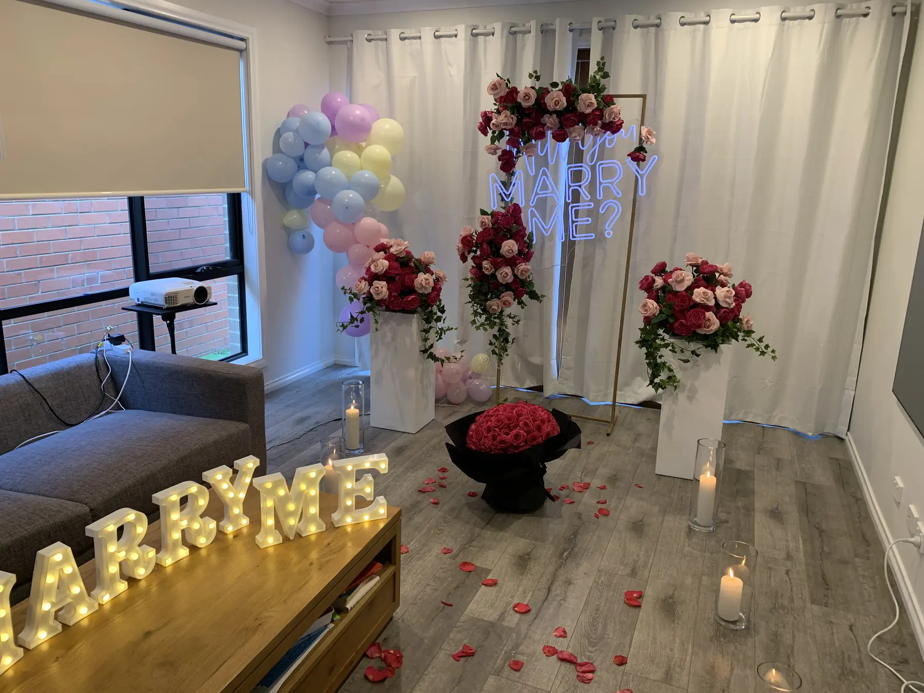 Full living room proposal setup with a pastel balloon garland, blush and burgundy florals on white pillars, a neon Marry Me sign, light-up MARRY ME letters, a rose bouquet and petals across the floor